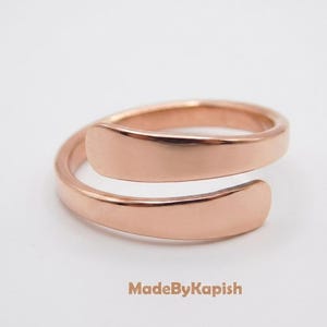 May include: A rose gold-toned bypass ring with a smooth, polished finish. The band features an open design where the two ends overlap slightly. The text MadeByKapish is printed in a light brown color at the bottom of the image.