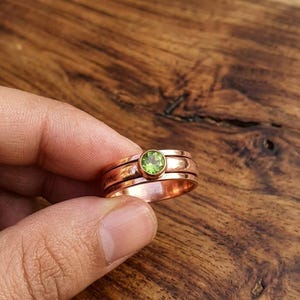 May include: A copper-colored ring with a green gemstone center. The ring has a spinning band design. The gemstone is round and set in a gold-colored bezel. The ring is displayed against a wooden background.