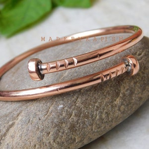 May include: A copper bangle bracelet with a hammered texture and two nail-shaped accents. The bracelet is open and can be adjusted to fit different wrist sizes.