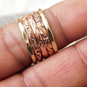 Puede incluir: Un conjunto de cinco anillos apilables de oro y cobre con diseños intrincados. Los anillos se llevan en un dedo.
