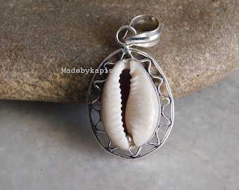 May include: A silver pendant featuring a natural cowrie shell encased in a decorative, openwork frame. The shell is off-white with a dark interior. The pendant has a loop for a chain and is displayed on a stone surface. The text "Madebykapish" is visible.