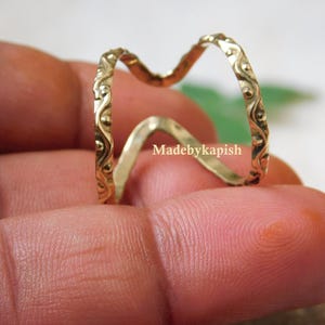 May include: A gold-colored ring with a unique double-wave design. The ring features an intricate, embossed pattern along its edges. The ring is held between fingers, showcasing its delicate details and craftsmanship. The text "Madebykapish" is visible.