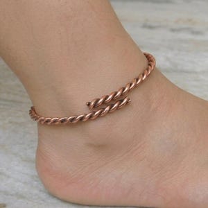 May include: A copper-colored, open-ended anklet with a twisted rope design. The anklet wraps around the ankle, with the ends slightly separated. The jewelry is a warm, reddish-brown color.