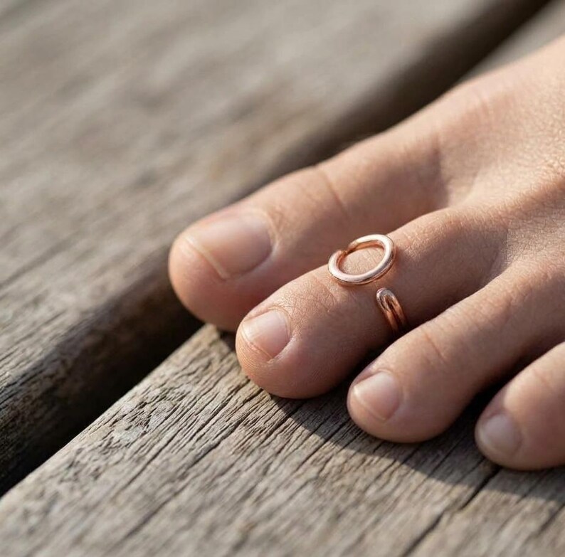 Op de afbeelding: Een open teenring in ros&eacute;goudkleur met een eenvoudig, glad, cirkelvormig ontwerp. De metalen ring wordt gedragen om de tweede teen en heeft een gepolijste afwerking die het licht reflecteert, met een kleine opening tussen de twee uiteinden.
