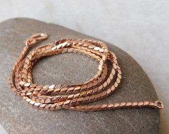 May include: A rose gold chain necklace with a delicate, flat, oval-shaped link design. The chain is coiled on a gray stone surface.