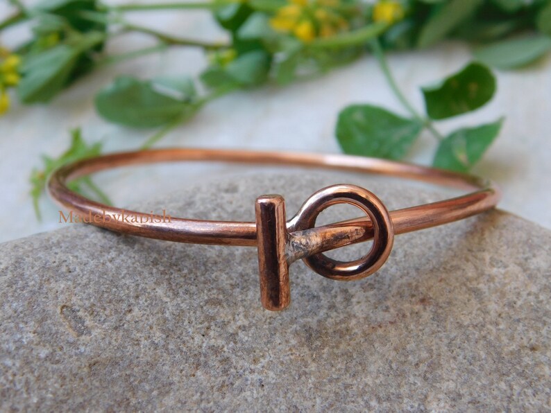 May include: A copper bangle bracelet with a simple, minimalist design. The bracelet features a single, rounded, copper clasp.