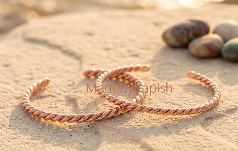 Puede incluir: Dos brazaletes de metal color cobre con un dise&ntilde;o de cuerda trenzada. Los brazaletes son abiertos y de forma circular. El texto MadeByKapish es visible en el centro de la imagen entre los dos brazaletes.