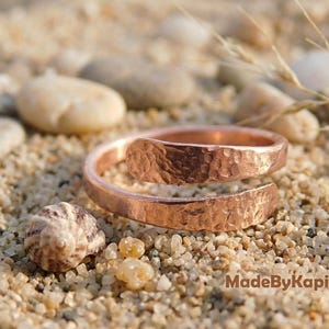 May include: A copper-colored bypass ring featuring a hammered, textured surface. The ring has an open, adjustable design with overlapping ends. The text MadeByKapish is visible in the lower right corner of the image, set against a sandy background.