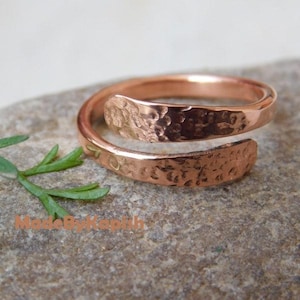 May include: A copper bypass ring featuring a hammered, textured finish on the outer surface. The band is open-ended, allowing for adjustable sizing, and displays a warm, metallic reddish-brown tone. The text MadeByKapish is visible.