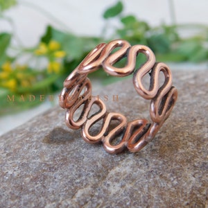 Può includere: Un anello color rame con un design ondulato unico. La forma dell'anello è composta da anelli allungati e interconnessi, creando un motivo continuo e fluido. L'anello è presentato su una superficie di pietra grigia strutturata. Il testo "MADE BY FISH" è visibile.