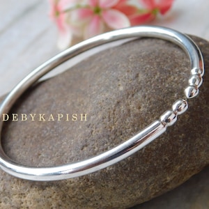 May include: A silver bangle bracelet with a row of small beads on one side. The bracelet is on a gray stone surface.