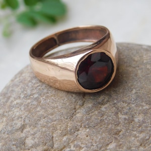 May include: A gold ring with a large, round, dark red gemstone set in the center. The ring has a thick band and a smooth, polished finish.