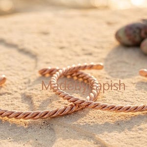 Puede incluir: Dos brazaletes de metal color cobre con un dise&ntilde;o de cuerda trenzada. Los brazaletes son abiertos y de forma circular. El texto MadeByKapish es visible en el centro de la imagen entre los dos brazaletes.
