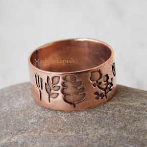 Op de afbeelding: Een koperen ring met een brede band met een gegraveerd ontwerp van bladeren, een vork en een bloem. De ring ligt op een bruine steen.