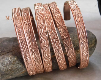 Può includere: Cinque braccialetti in rame con motivi floreali. I braccialetti sono impilati uno sopra l'altro e hanno una finitura lucida e lucidata.
