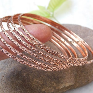 May include: Set of seven copper bangles with an intricate, repeating pattern. The bangles are stacked on top of each other and have a warm, metallic sheen.