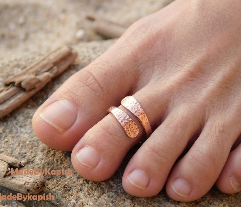 May include: A copper-colored toe ring featuring a textured, hammered finish. The ring has an open, bypass-style design with two tapered ends that wrap slightly around the toe. The surface of the metal has a distinct, dimpled appearance.