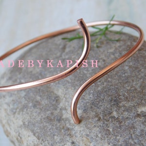 May include: A simple copper bangle bracelet with a curved design. The bracelet is made of a single piece of wire that is bent into a circle and then twisted at one end to create a unique shape.