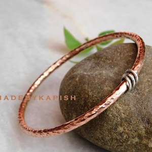 Könnte beinhalten: Ein gehämmertes Kupferarmband mit einem silbernen Akzent. Das Armband liegt auf einem braunen Stein.