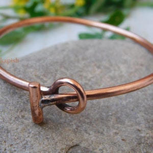 May include: A copper bangle bracelet with a simple knot design. The bracelet is made of a single piece of copper wire that is bent into a circle and then twisted to create the knot. The bracelet is a simple and elegant piece of jewelry that can be worn on its own or stacked with other bracelets.