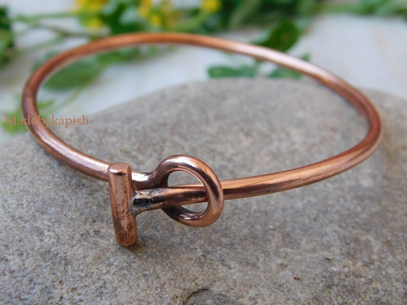 May include: A copper bangle bracelet with a simple knot design. The bracelet is made of a single piece of copper wire that is bent into a circle and then twisted to create the knot. The bracelet is a simple and elegant piece of jewelry that can be worn on its own or stacked with other bracelets.