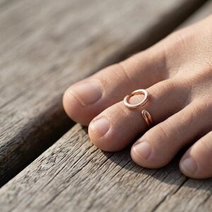 Op de afbeelding: Een open teenring in ros&eacute;goudkleur met een eenvoudig, glad, cirkelvormig ontwerp. De metalen ring wordt gedragen om de tweede teen en heeft een gepolijste afwerking die het licht reflecteert, met een kleine opening tussen de twee uiteinden.