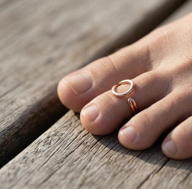 Op de afbeelding: Een open teenring in ros&eacute;goudkleur met een eenvoudig, glad, cirkelvormig ontwerp. De metalen ring wordt gedragen om de tweede teen en heeft een gepolijste afwerking die het licht reflecteert, met een kleine opening tussen de twee uiteinden.