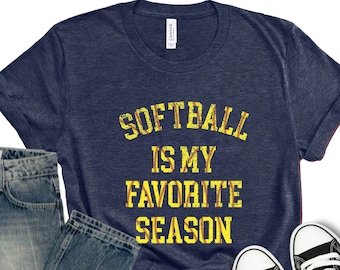 May include: A navy blue t-shirt with the text "Softball is my favorite season" in yellow. The shirt is paired with blue jeans and black Converse sneakers.