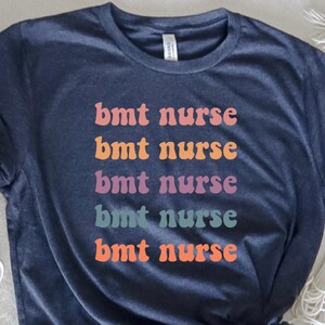 May include: A dark blue t-shirt with the text "bmt nurse" repeated five times in different colors. The colors are pink, purple, teal, orange, and peach.