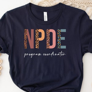 May include: A navy blue t-shirt with the text "NPDE program coordinator" in a colorful, patterned font. The letters are decorated with a leopard print design.