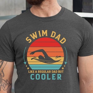 May include: A dark heather grey t-shirt with a vintage-style graphic of a swimmer in a pool. The text "Swim Dad Like a Regular Dad But Cooler" is printed on the shirt.