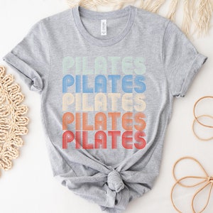 May include: A gray t-shirt with the word "PILATES" repeated five times in a retro style font. Each word is a different color, starting with light green, then blue, yellow, orange, and red.