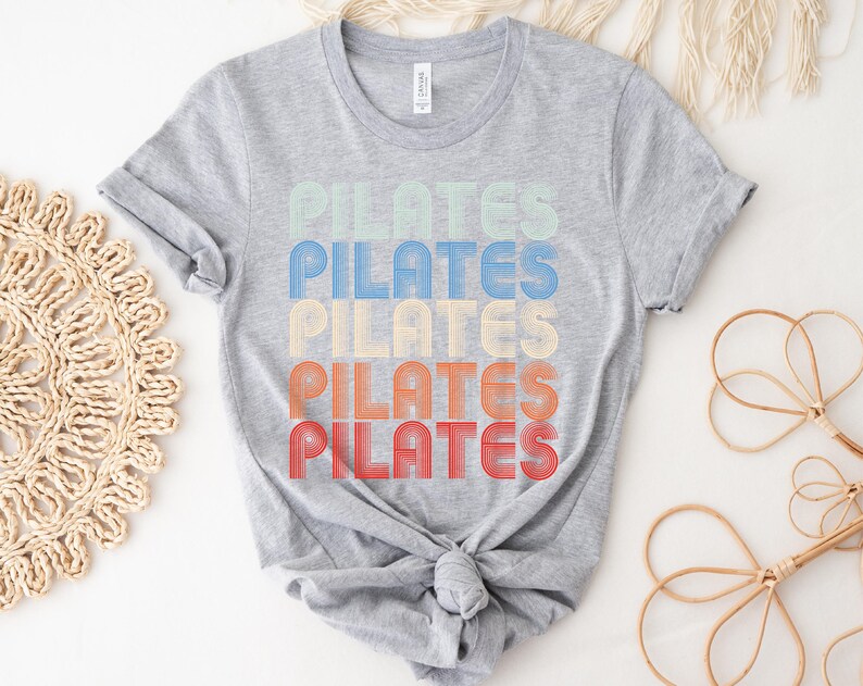 May include: A gray t-shirt with the word "PILATES" repeated five times in a retro style font. Each word is a different color, starting with light green, then blue, yellow, orange, and red.