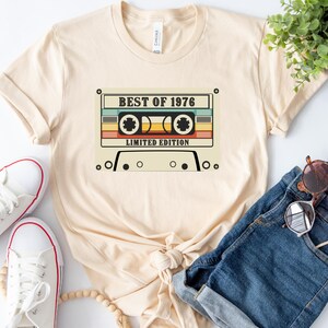May include: A beige t-shirt with a vintage cassette tape graphic. The tape has the text "Best of 1976 Limited Edition" printed on it.