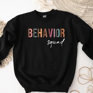 May include: Black sweatshirt with the text "BEHAVIOR squad" in colorful letters with a leopard print pattern.