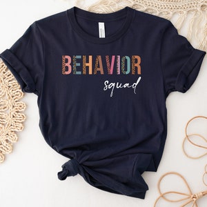 May include: A navy blue t-shirt with the text "BEHAVIOR squad" in colorful letters with a patterned design.