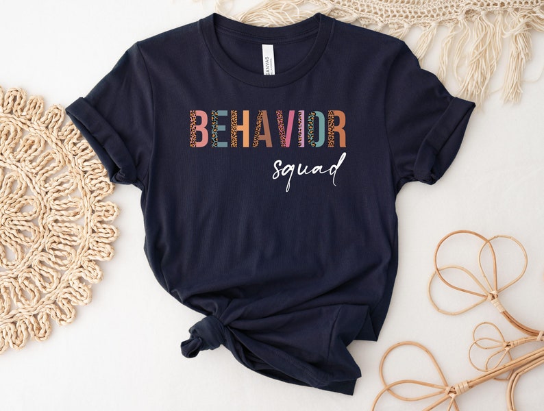 May include: A navy blue t-shirt with the text "BEHAVIOR squad" in colorful letters with a patterned design.