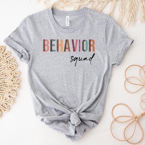 May include: A gray t-shirt with the text "BEHAVIOR squad" printed on the front. The letters are in a colorful, patterned font.