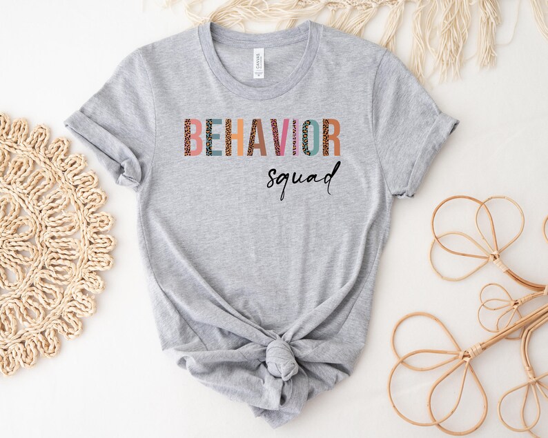 May include: A gray t-shirt with the text "BEHAVIOR squad" printed on the front. The letters are in a colorful, patterned font.