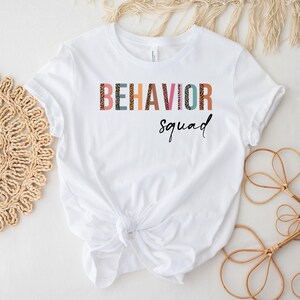 May include: White t-shirt with the text "BEHAVIOR squad" in colorful letters with a leopard print pattern.