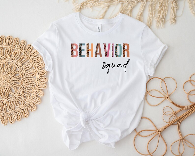 May include: White t-shirt with the text "BEHAVIOR squad" in colorful letters with a leopard print pattern.