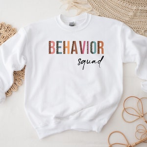 May include: White sweatshirt with the text "BEHAVIOR squad" in colorful letters with a leopard print pattern.