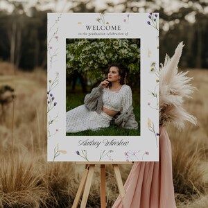 May include: A white welcome sign with a floral border and a photo of a woman in a white dress and a gray cardigan. The sign says "Welcome to the graduation celebration of Audrey Winslow".