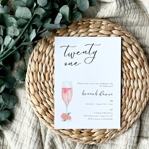 May include: A white invitation card with black calligraphy that reads "Twenty One". The card features a watercolor illustration of a glass of pink champagne with pink roses at the base. The text on the card reads "Please join us as we celebrate the birthday girl Hannah Davis on Saturday, August 15th at A Splash of Red Winery, 2155 Juniper Love, Sugar City, MT. RSVP to Jessica at 123-456-7890."