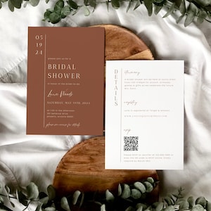 May include: Two bridal shower invitation cards. The main card is a warm brown with the text "BRIDAL SHOWER" and event details. The second card is white with event details and a QR code. The cards are on a wooden surface.