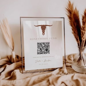 May include: A framed sign with a brown bull skull graphic and the words "HONEYMOON FUND". A QR code is below the text, with the names "Sadie + Hudson" and a date. The sign is displayed on a beige surface with decorative elements.