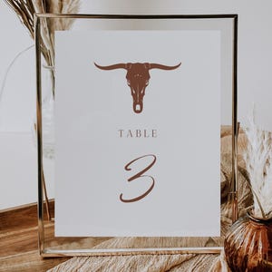 May include: A gold framed table number sign with a brown bull skull graphic and the text "TABLE 3" in a brown script font.