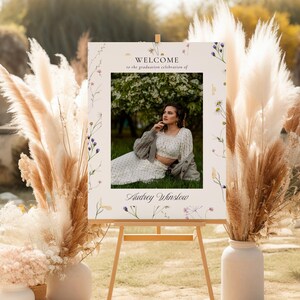 May include: A welcome sign for a graduation celebration with a floral design and a photo of a person in a white dress and a cardigan. The sign says "Welcome to the graduation celebration of Audrey Winslow".