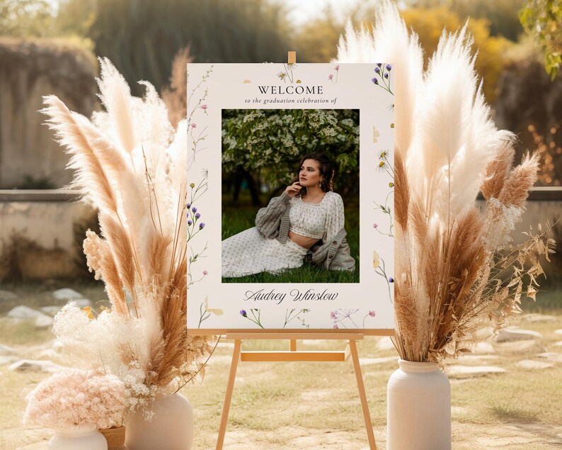 May include: A welcome sign for a graduation celebration with a floral design and a photo of a person in a white dress and a cardigan. The sign says "Welcome to the graduation celebration of Audrey Winslow".