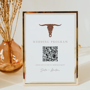 May include: A white sign with a gold frame, featuring a brown bull skull graphic and a QR code. The text "WEDDING PROGRAM" is printed in brown above the QR code. Below the QR code, the text "please scan to view our wedding program" is printed in brown. The names "Sadie + Hudson" are printed in brown cursive below the text.